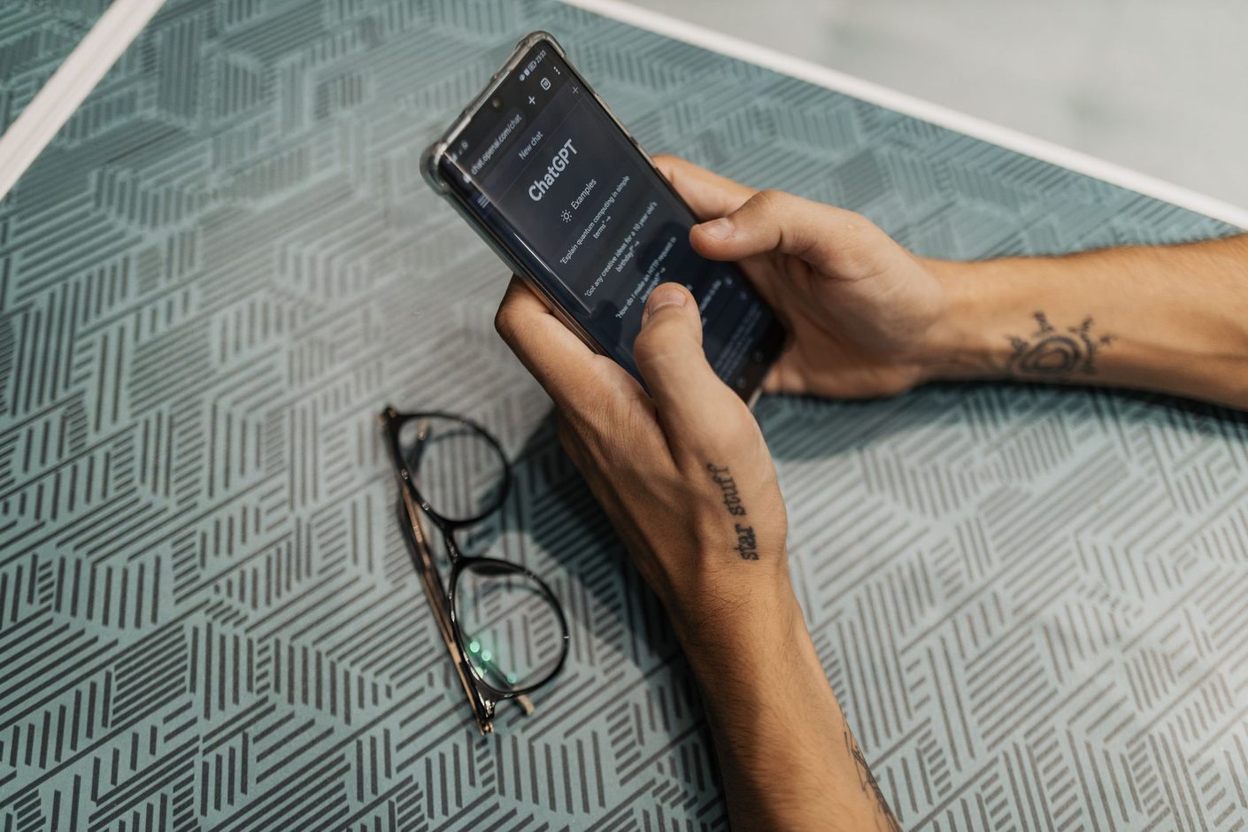Two hands holding a smartphone with the ChatGPT app open, a pair of glasses lying on the table beside it