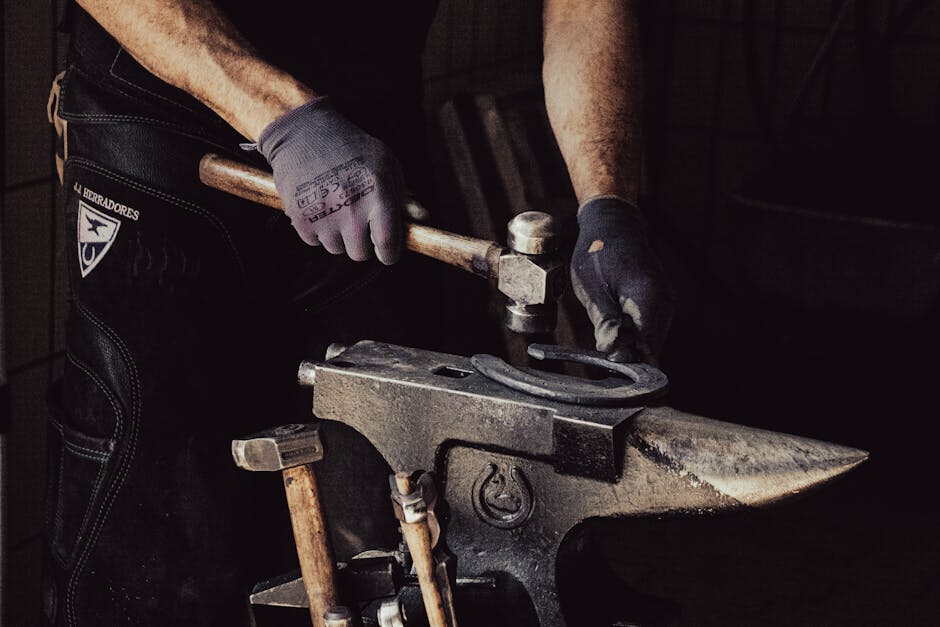 Hands in work gloves forging a horseshoe with a hammer on an anvil in a dark workshop
