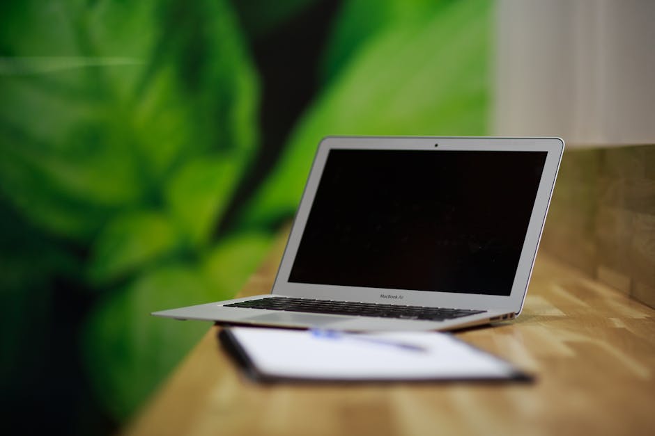 Open silver laptop on a wooden table with a clipboard and pen in front of it, blurred greenery in the background